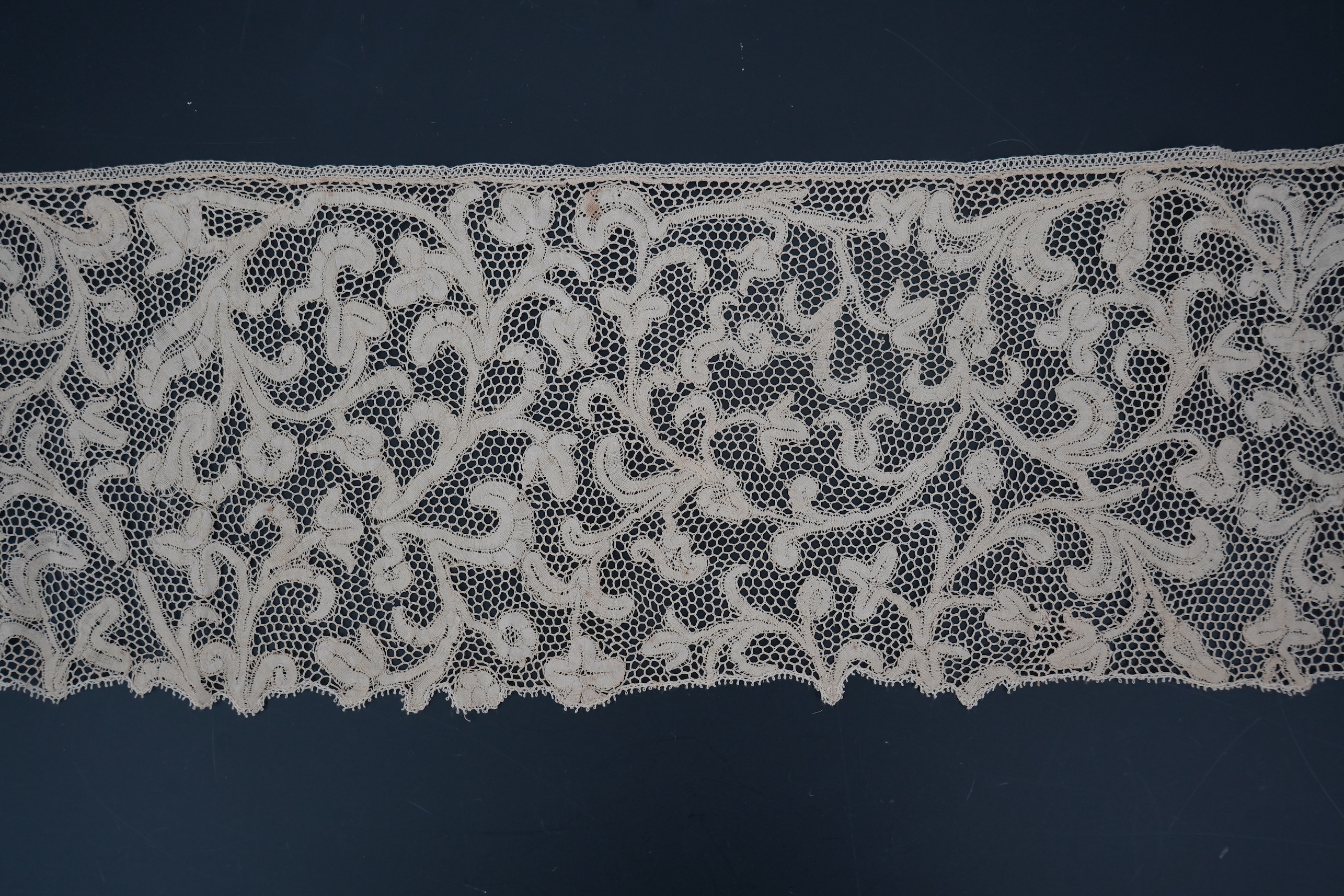 A length of 1740-1760 Italian, Milanese bobbin lace flounce, made from flat braiding or tapes, placed into scrolling patterns, attached to brides, made into a honeycomb mesh. 252cm long x 16cm deep. Condition - one small